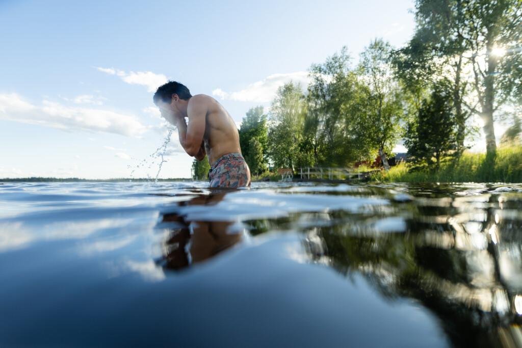 Mies menossa uimaan. Samalla hän kastaa kasvojaan veteen. Taustalla sininen taivas ja vihreitä puita, joiden takaa paistaa aurinko.