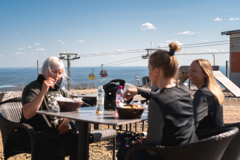 Three people eating and taking a break outdoors at the top of Yllästunturi at Ylläskammi. The day is sunny. In the background, the Ylläs gondola lift is clearly visible, along with an open landscape on the horizon and a blue sky.