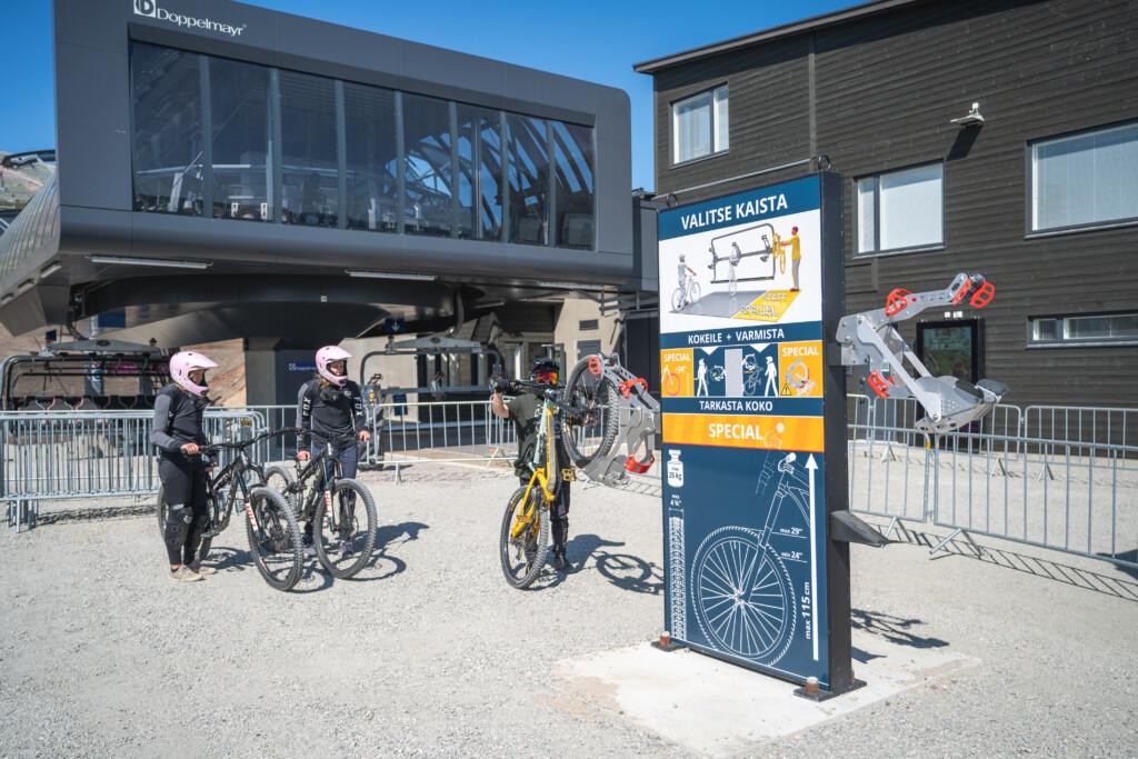Three riders practicing how to load a bike onto the chairlift at Ylläs Bike Park. A training device is in the foreground, with the chairlift building and riders in the background.