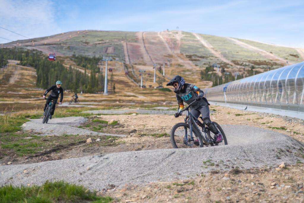 Three riders descending the Ylläs Bike Park training track next to the Vekkuli magic carpet lift. In the background, Yllästunturi fell, the gondola lift, and a blue sky.