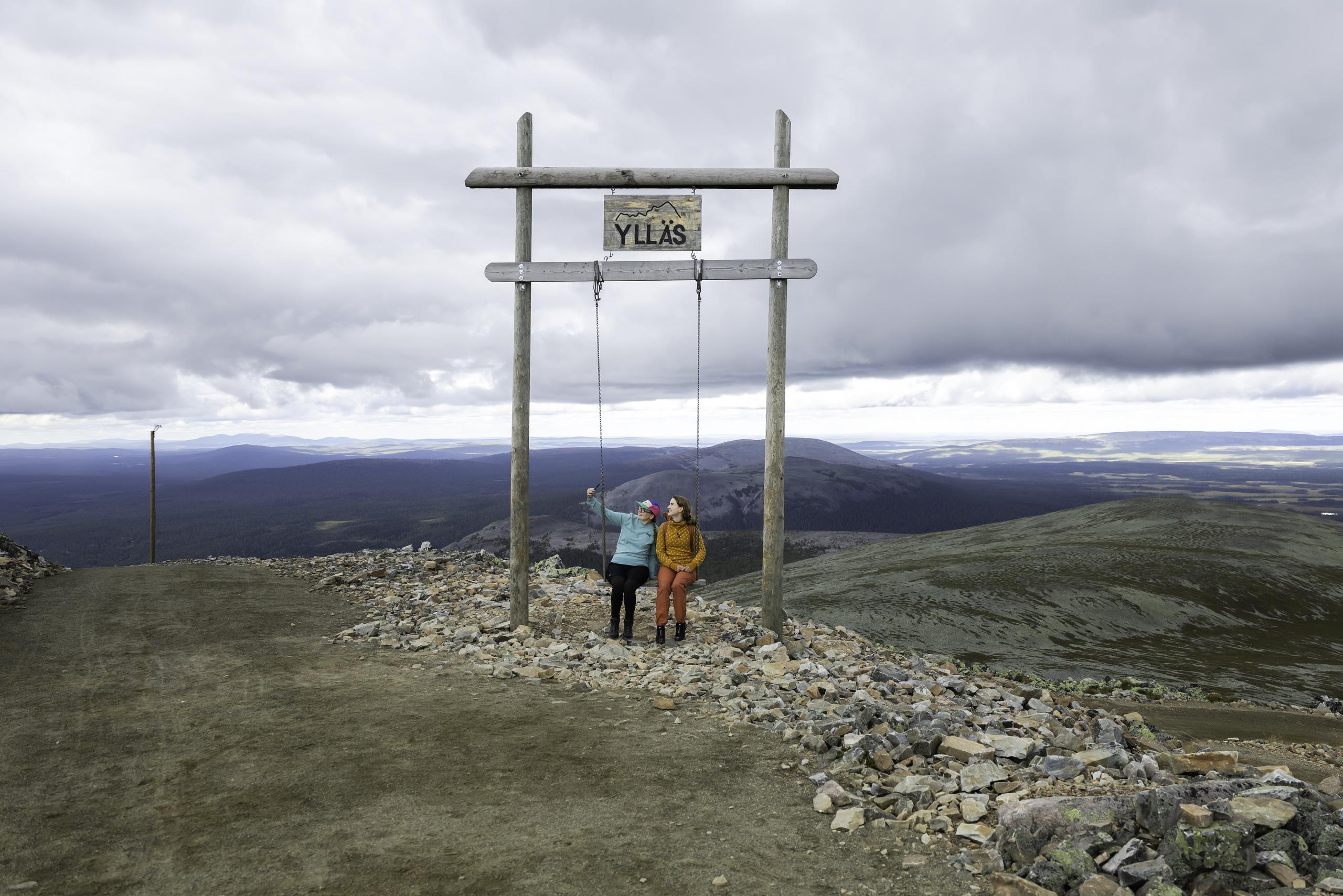 Two people sitting on the large swing at the top of Yllästunturi. They are taking a selfie. In the background, there is a fell landscape and a cloudy sky.