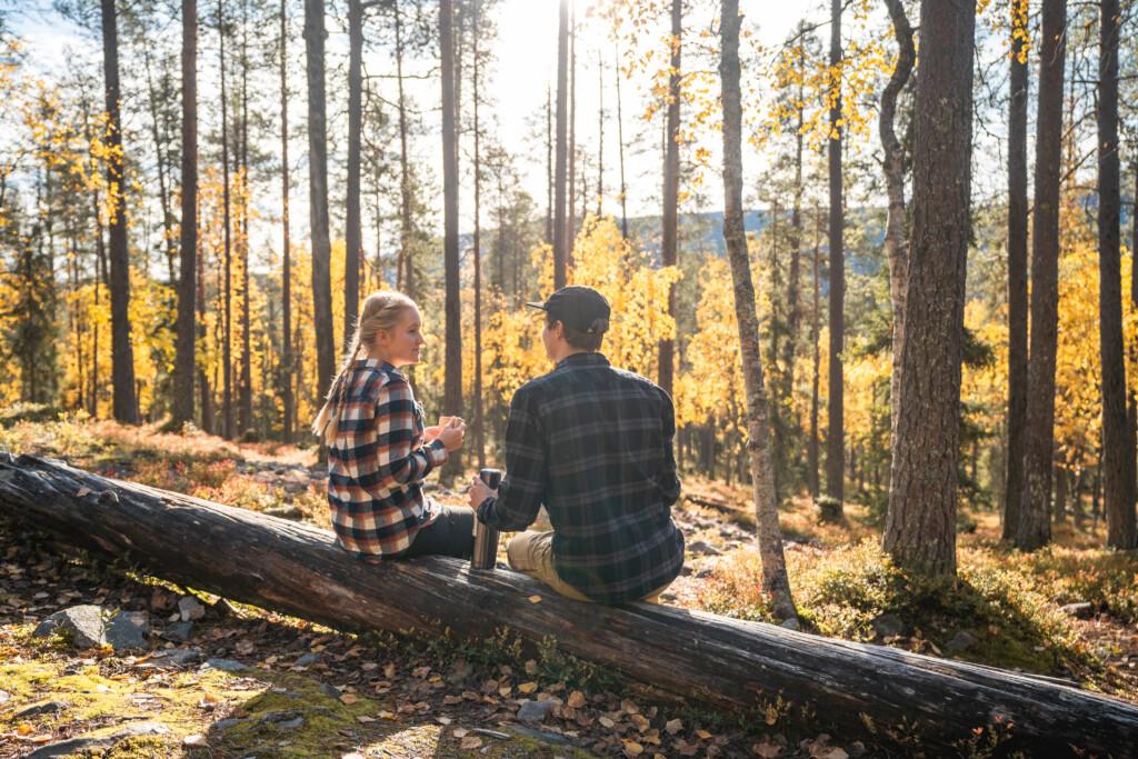 Two people sitting on a fallen tree trunk, holding a thermos flask. Sunlight filters through autumn-coloured trees into the forest.
