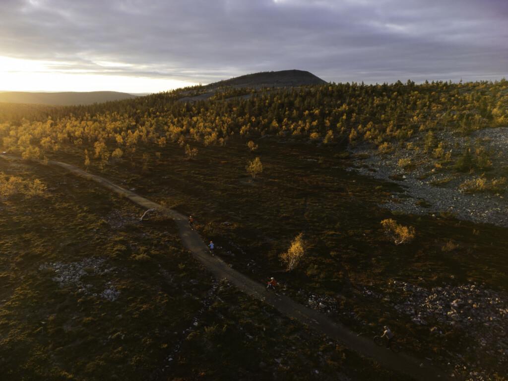 Kolme pyöräilijää ajaa polulla ylhäällä tunturissa. Puissa on ja ympäristössä on ruskan värit. Taustalla tunturin huippu ja matalalta paistava aurinko.