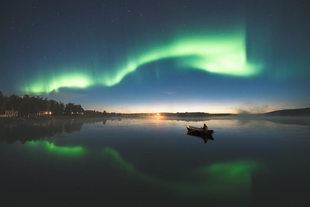 Etualalla ihminen järvellä soutuveneessä, taivaalla loimuaa isot vihertävät revontulet ja tuikkii tähtiä. Rannalla näkyy valoja ja muutamia rakennuksia.