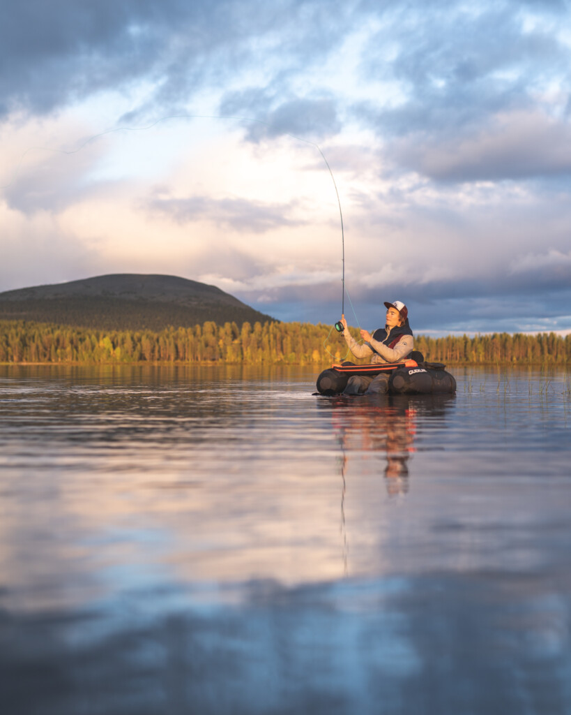Mies kalastaa renkaalla kelluen Äkäslompolojärven selällä. Taustalla Kesänkitunturi ja hieman pilvinen taivas.