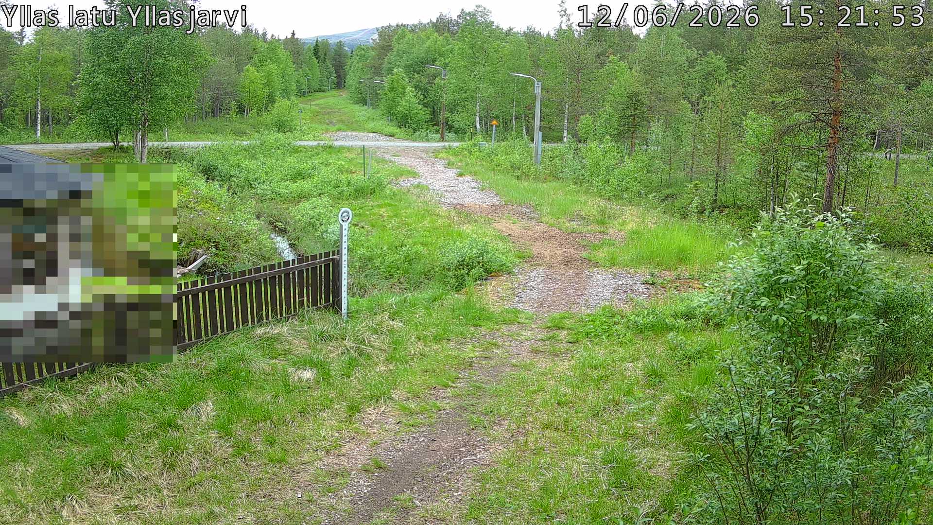 Talvella hieman mutkitteleva hiihtolatu, kesällä metsää, ja kauempana taustalla puiden yläpuolella hieman Yllästunturin rinnettä.