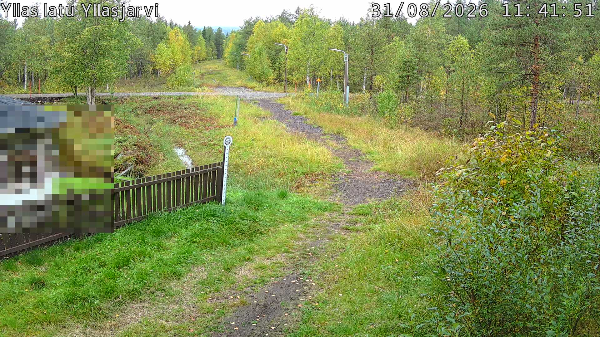 Talvella hieman mutkitteleva hiihtolatu, kesällä metsää, ja kauempana taustalla puiden yläpuolella hieman Yllästunturin rinnettä.
