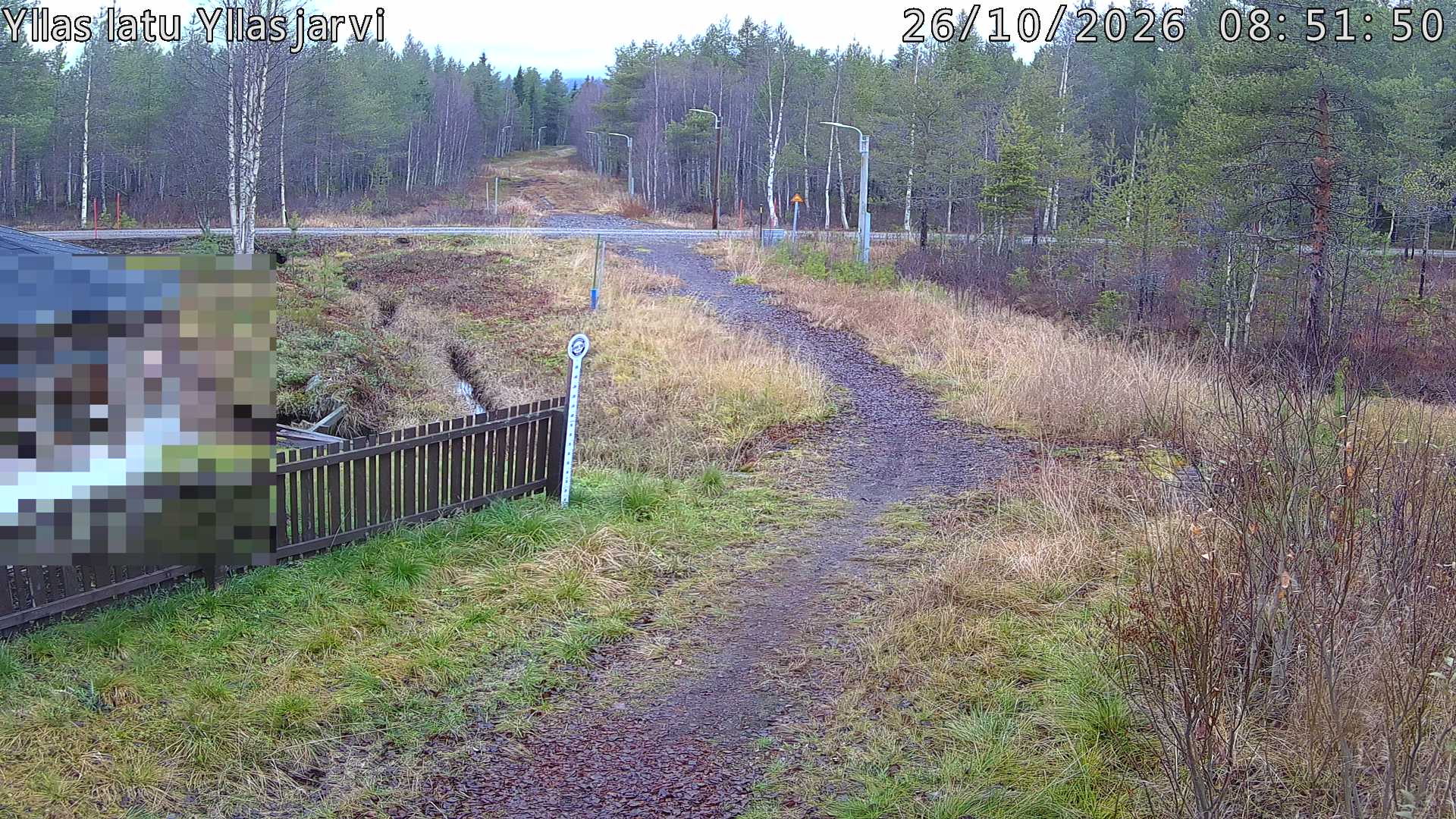 Talvella hieman mutkitteleva hiihtolatu, kesällä metsää, ja kauempana taustalla puiden yläpuolella hieman Yllästunturin rinnettä.