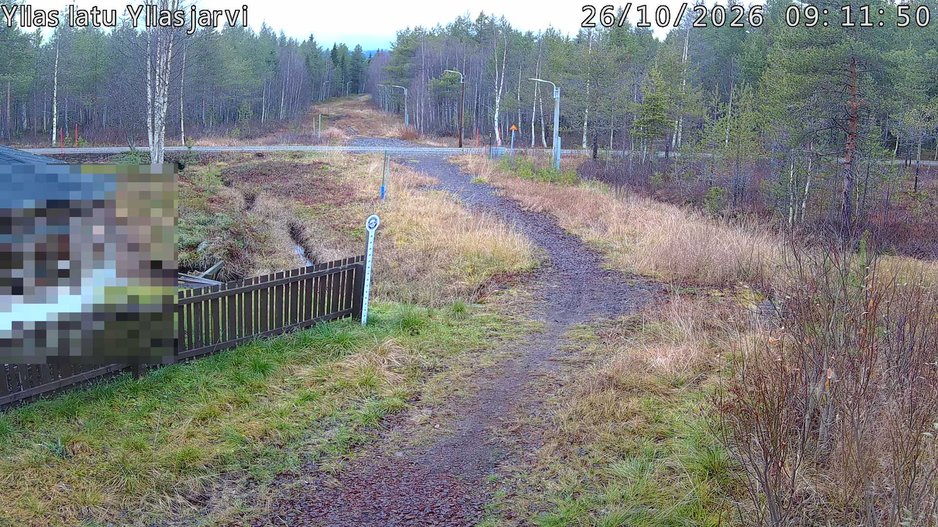 Talvella hieman mutkitteleva hiihtolatu, kesällä metsää, ja kauempana taustalla puiden yläpuolella hieman Yllästunturin rinnettä.