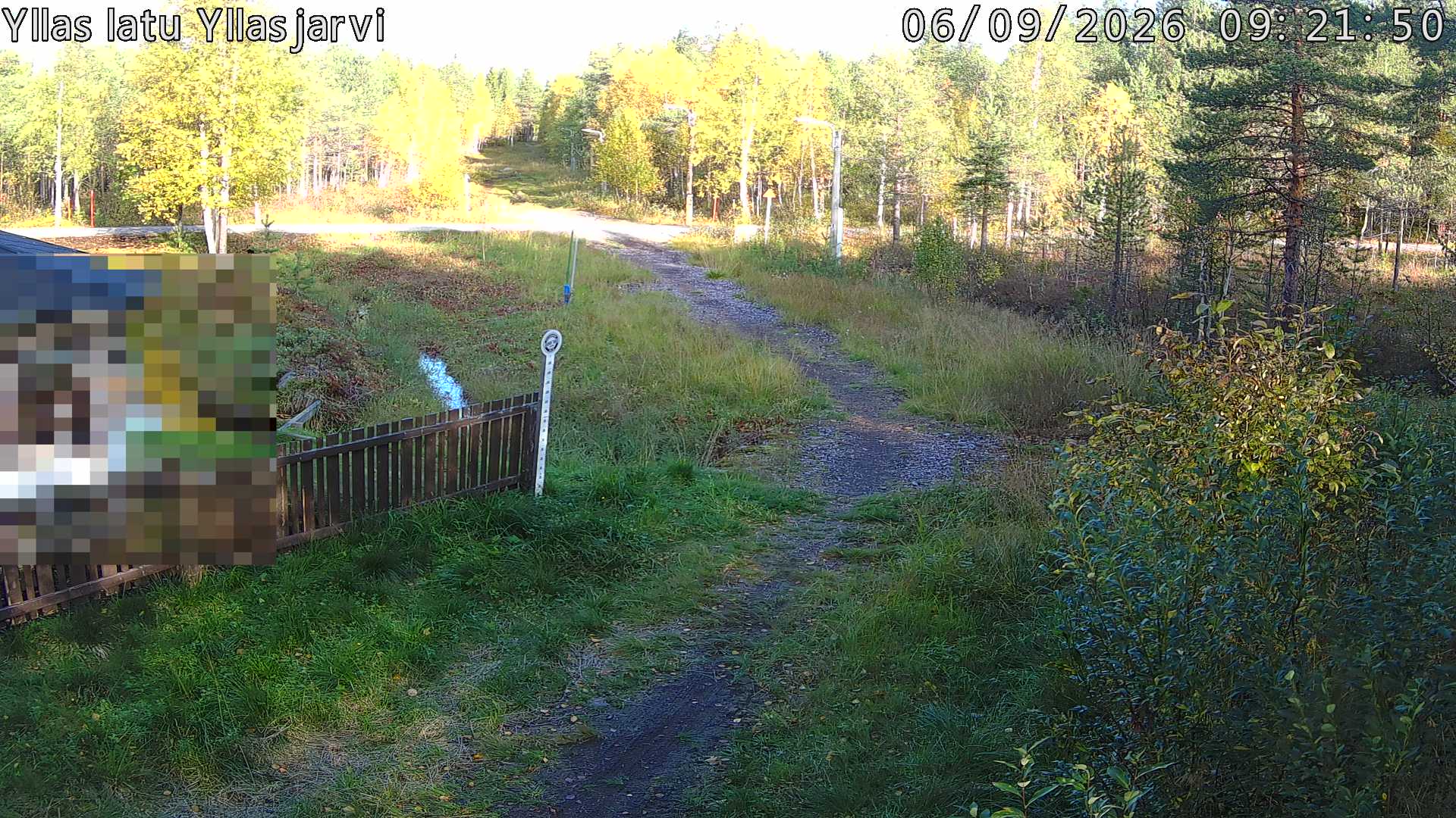 Talvella hieman mutkitteleva hiihtolatu, kesällä metsää, ja kauempana taustalla puiden yläpuolella hieman Yllästunturin rinnettä.