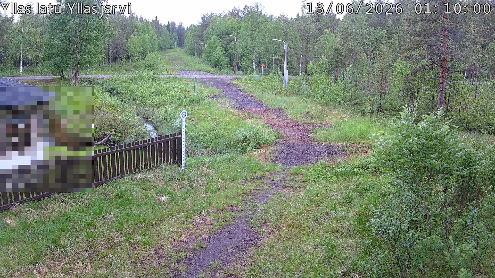 Talvella hieman mutkitteleva hiihtolatu, kesällä metsää, ja kauempana taustalla puiden yläpuolella hieman Yllästunturin rinnettä.