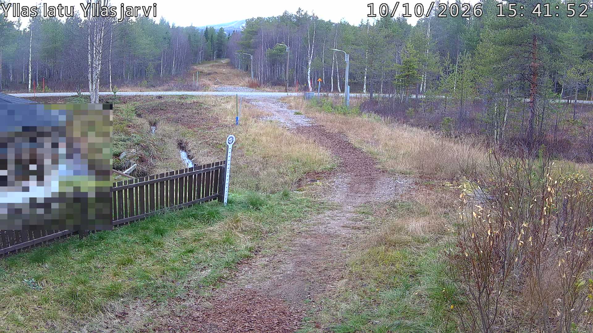 Talvella hieman mutkitteleva hiihtolatu, kesällä metsää, ja kauempana taustalla puiden yläpuolella hieman Yllästunturin rinnettä.