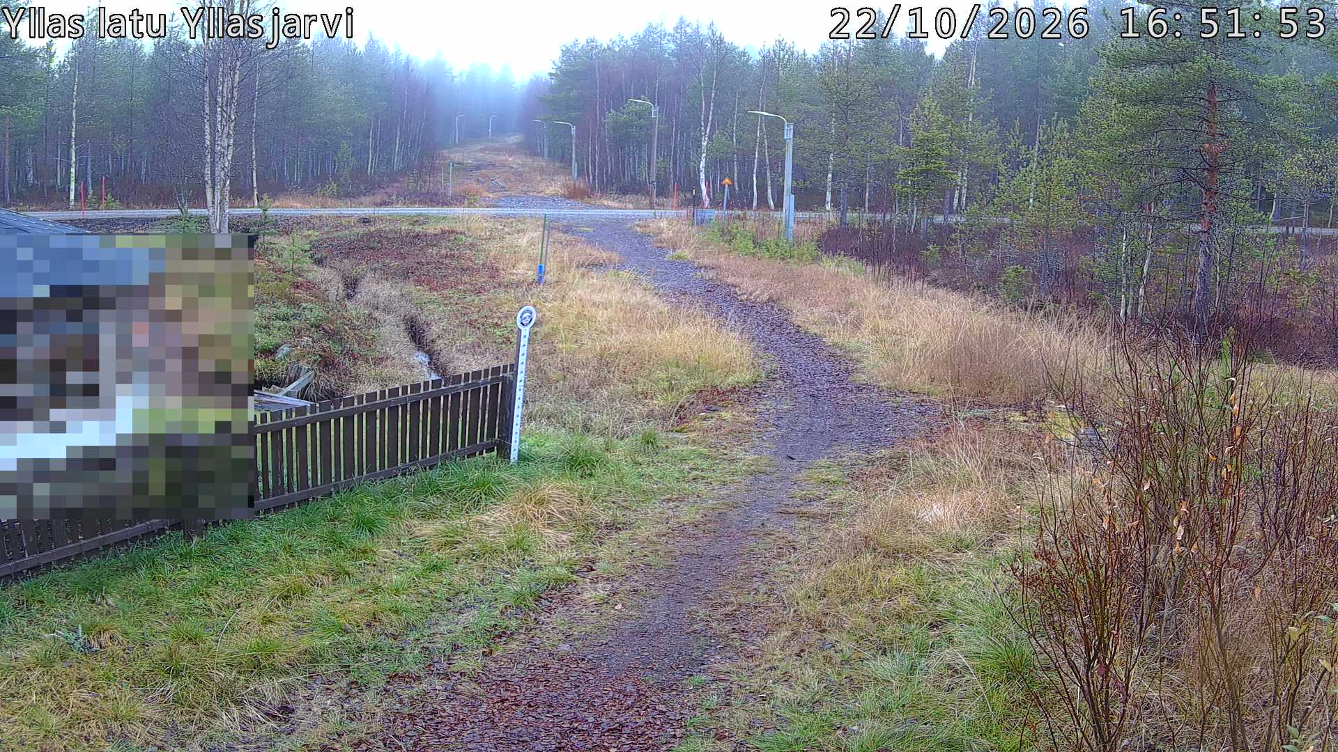 Talvella hieman mutkitteleva hiihtolatu, kesällä metsää, ja kauempana taustalla puiden yläpuolella hieman Yllästunturin rinnettä.
