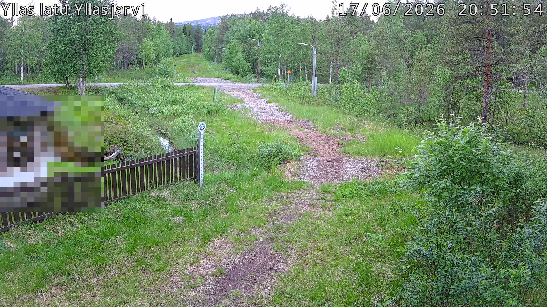 Talvella hieman mutkitteleva hiihtolatu, kesällä metsää, ja kauempana taustalla puiden yläpuolella hieman Yllästunturin rinnettä.