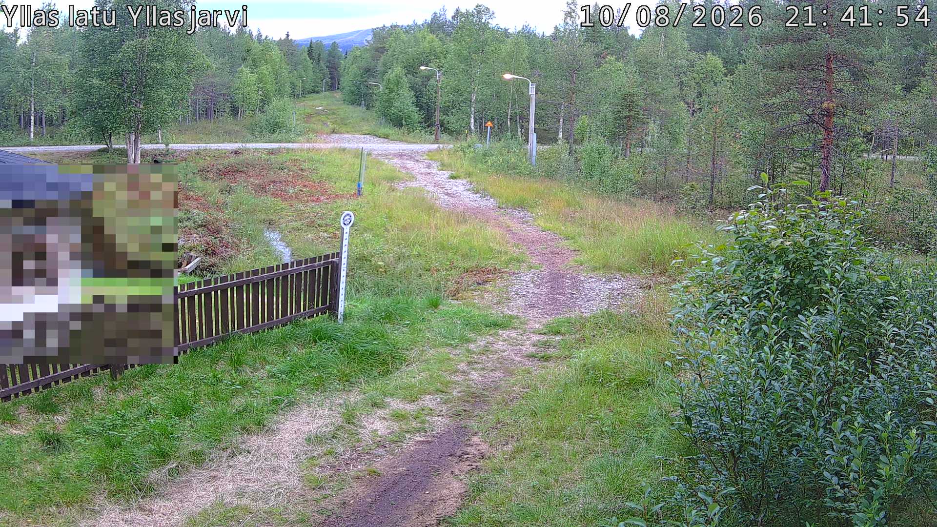 Talvella hieman mutkitteleva hiihtolatu, kesällä metsää, ja kauempana taustalla puiden yläpuolella hieman Yllästunturin rinnettä.