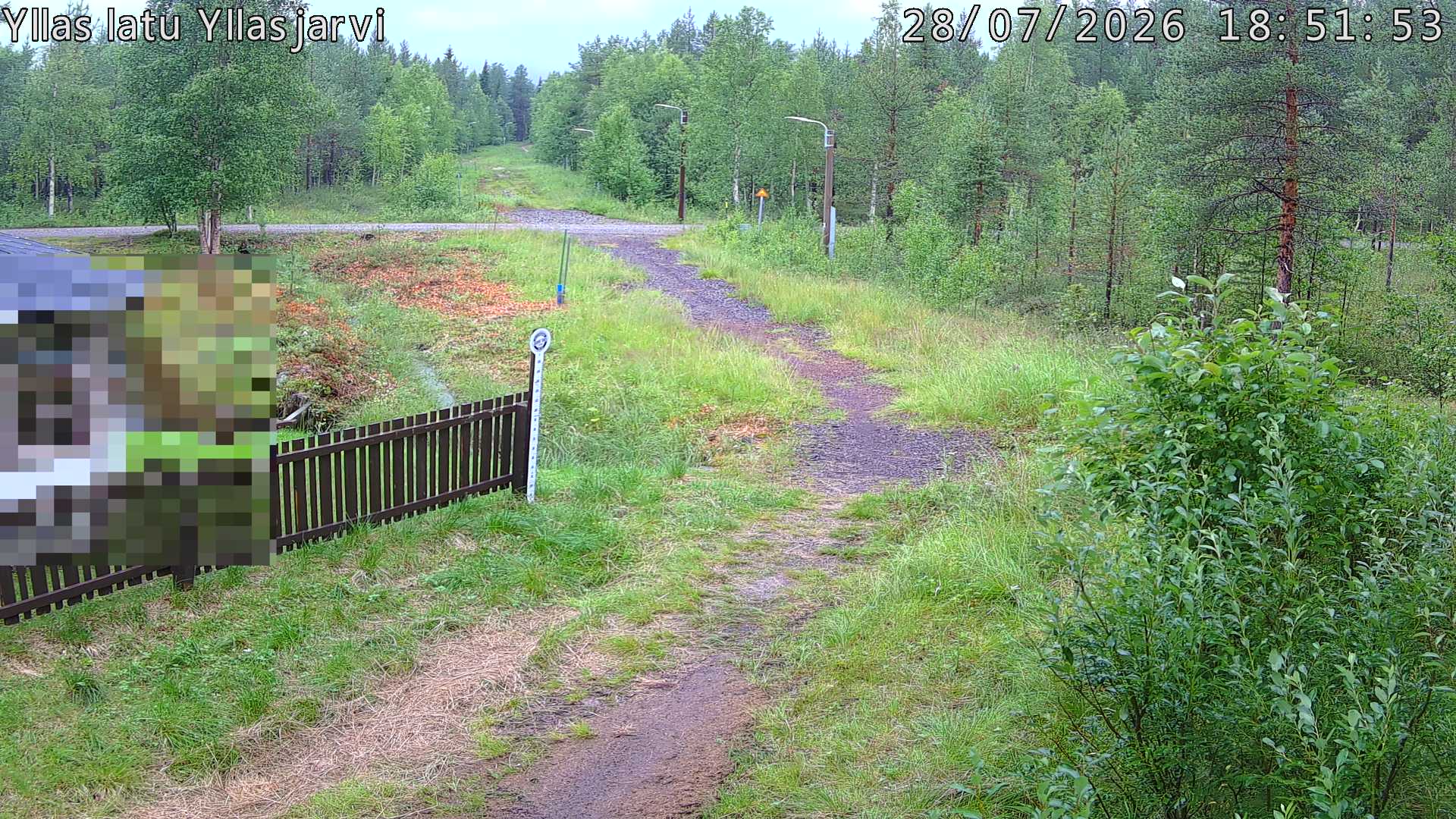 Talvella hieman mutkitteleva hiihtolatu, kesällä metsää, ja kauempana taustalla puiden yläpuolella hieman Yllästunturin rinnettä.