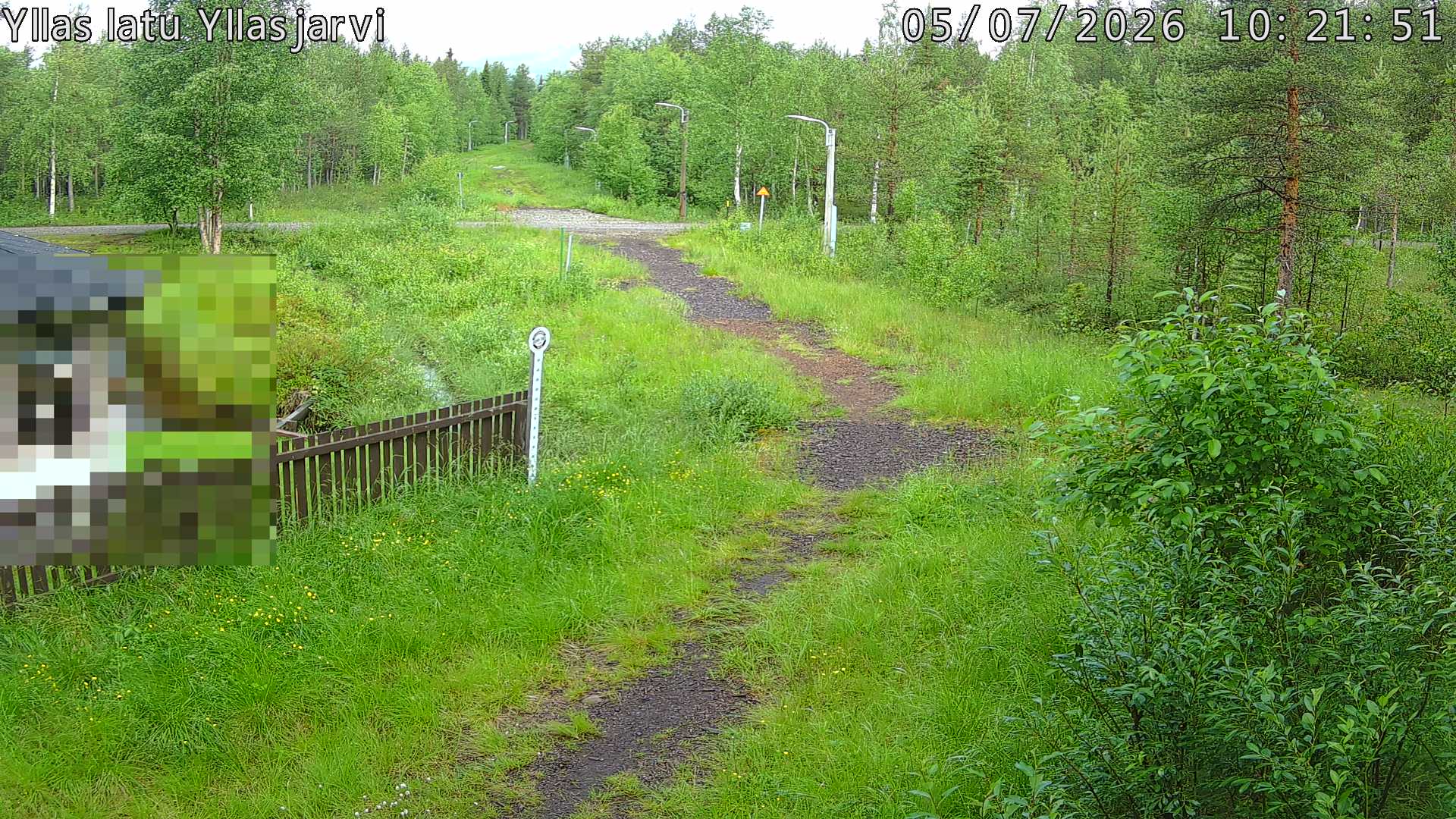 Talvella hieman mutkitteleva hiihtolatu, kesällä metsää, ja kauempana taustalla puiden yläpuolella hieman Yllästunturin rinnettä.